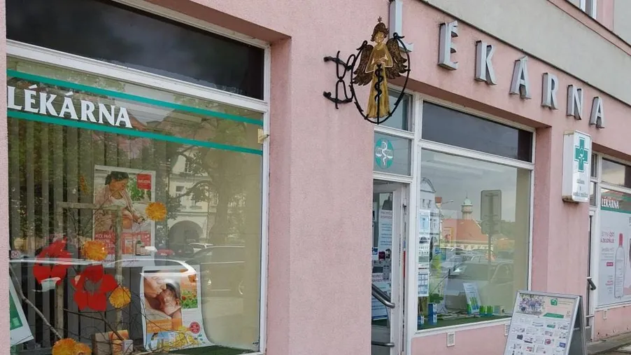 Vchod do lékárny v růžové budově s výlohou, nápisem "LÉKÁRNA" a tabulí s informacemi před vstupem.